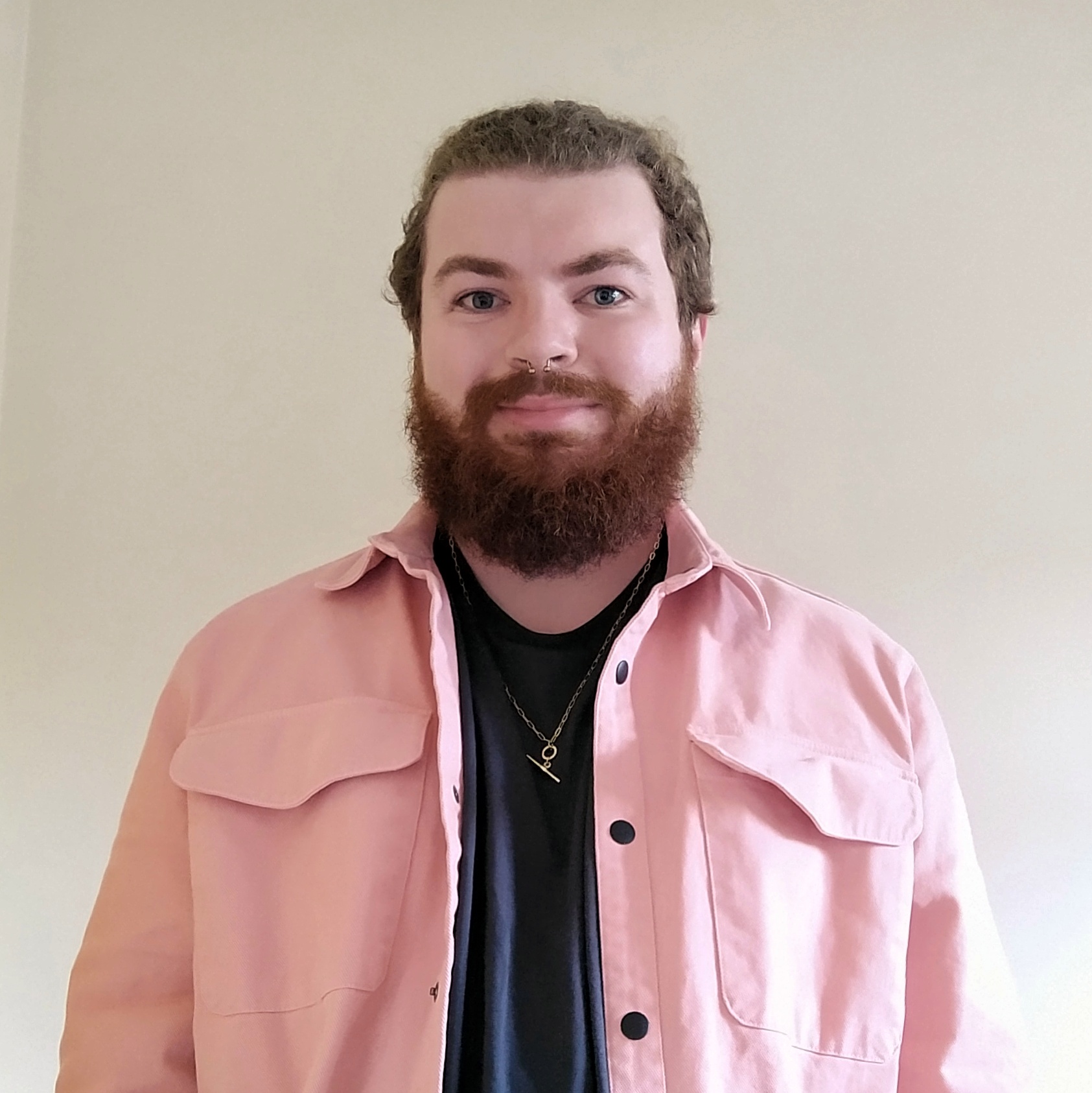 My headshot. I am smiling slightly, with my light brown curly hair pulled back into a ponytail, and my reddish beard a medium length. I have a gold horseshoe septum piercing. I am wearing a grey-black tank top under a dusty pink shirt-jacket with black buttons, and a thin gold chain with a T-bar. The background is plain cream.